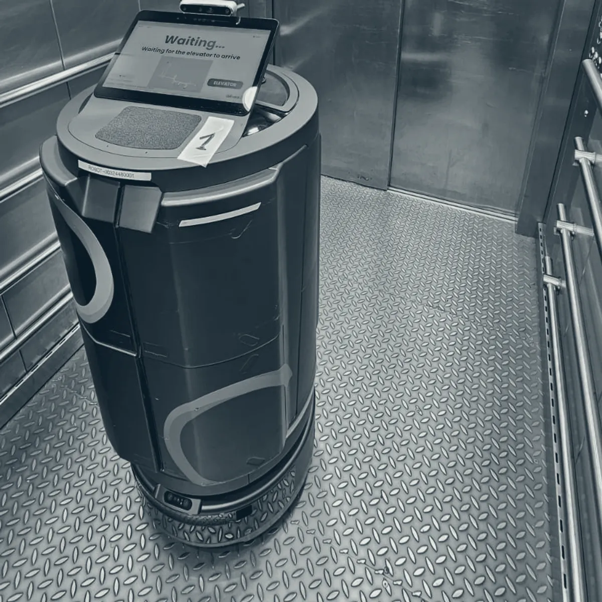 A delivery robot alone inside a service lift, its screen reading that it is waiting for the lift to arrive.